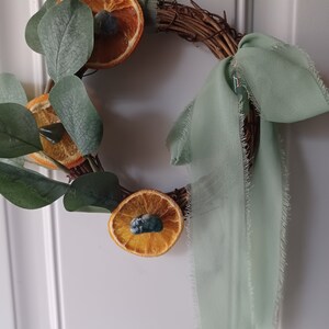 May include: A green fabric ribbon tied around a grapevine wreath with dried orange slices and eucalyptus leaves.