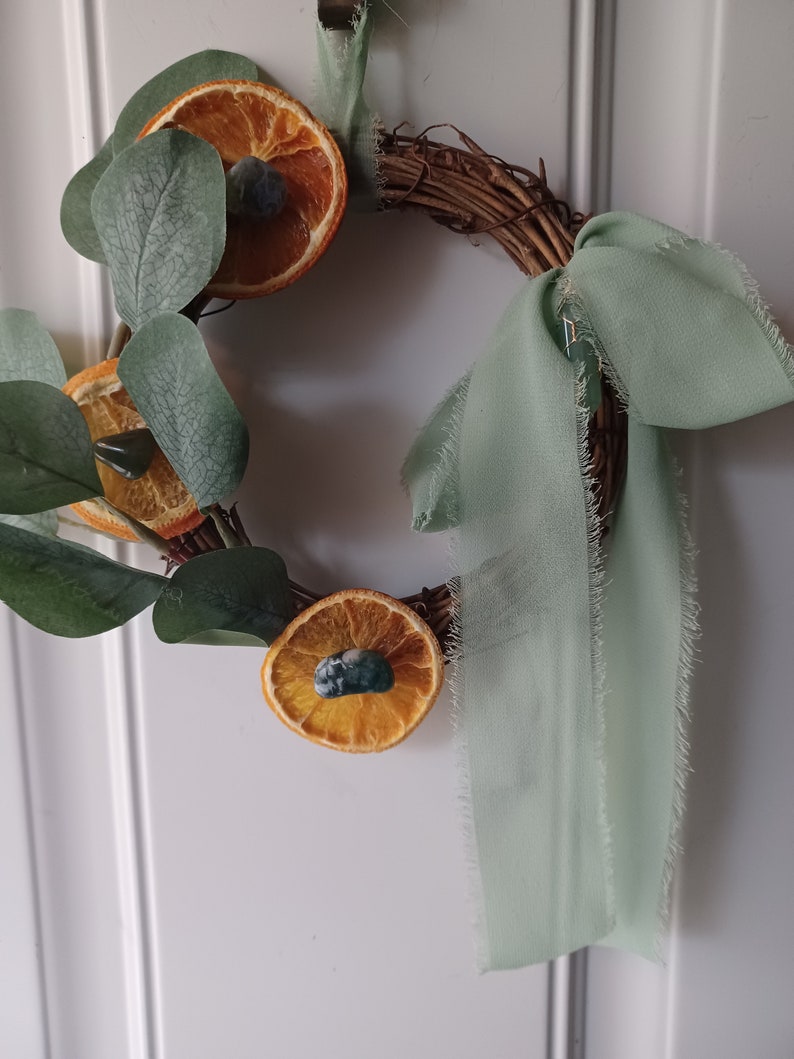 May include: A green fabric ribbon tied around a grapevine wreath with dried orange slices and eucalyptus leaves.