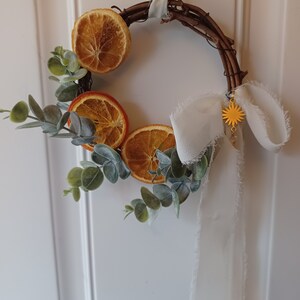 May include: A small, rustic wreath made of twigs with dried orange slices and eucalyptus leaves. A white ribbon with frayed edges hangs from the wreath, and a gold star charm dangles from the ribbon.