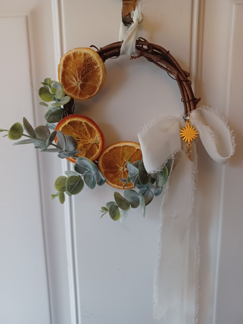 May include: A small, rustic wreath made of twigs with dried orange slices and eucalyptus leaves. A white ribbon with frayed edges hangs from the wreath, and a gold star charm dangles from the ribbon.