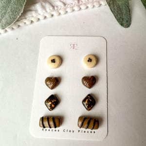 Puede incluir: Un conjunto de seis pendientes de arcilla con forma de caramelos de chocolate. Los pendientes están en una tarjeta blanca con el texto "Reeces Clay Pieces".