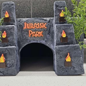 May include: A grey, rock-textured animal hideout featuring a central arched entrance and the text JURASSK PARK in orange lettering. The structure is adorned with six small, orange and yellow flame accents and green artificial fern foliage.