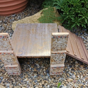 May include: A wooden model of a bridge with decorative carvings on the sides. The bridge is made of brown wood and has a light brown finish. The bridge is sitting on a bed of small stones.