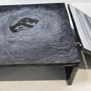 May include: A black wooden box with a dinosaur silhouette on the lid. The box is open and has a grey interior with multiple compartments.