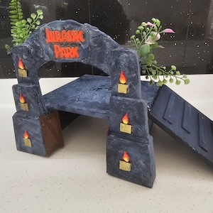 May include: A grey stone archway with the words "Jurassic Park" in red letters. The archway has a small platform and a ramp leading up to it. There are small orange flames on the sides of the archway.