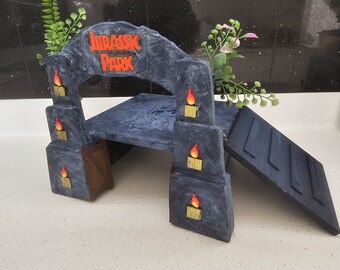 May include: A gray stone archway with the words "Jurassic Park" in red letters. The archway has a small platform and a ramp leading up to it. There are small orange flames on the sides of the archway.