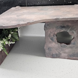 May include: A stone-effect decorative structure with a naturalistic design. It has a tabletop supported by a textured base with a cave-like opening. Artificial greenery and white flowers are placed within a recessed area.