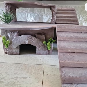 May include: A multi-level, brown faux-stone structure with a cave-like opening, stairs, and a ramp. The structure has a flat top with a plant and is decorated with small green plants. The design is suitable for small pets.