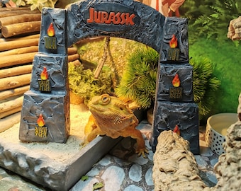 May include: A bearded dragon lizard in a terrarium with a Jurassic Park themed archway. The arch is gray with the word "JURASSIC" in red. The background features a forest scene and a picture of Mr. Bean.