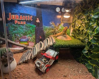 May include: A terrarium with a Jurassic Park theme. A tan lizard rests on a grassy platform. A toy Jeep and a wooden bridge are in the foreground. The background features a Jurassic Park sign and a jungle backdrop.