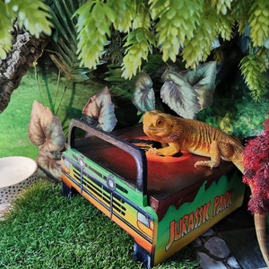 May include: A yellow and green toy truck with the words "Jurassic Park" printed on the side. A bearded dragon lizard is sitting on top of the truck.