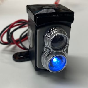 Puede incluir: Una luz con forma de cámara negra y plateada con una luz LED azul. La luz está cableada con cables rojos y negros.