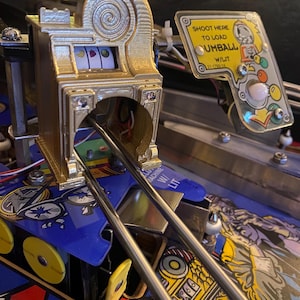 Könnte beinhalten: Nahaufnahme eines Flipperautomaten mit einem goldfarbenen Spielautomaten-Element. Der Text "SHOOT HERE TO LOAD GUMBALL" ist sichtbar. Die Maschine hat ein blaues und gelbes Design mit silbernen Metallstangen.