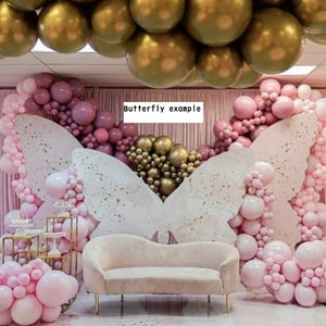 Op de afbeelding: Een roze en gouden ballonboog met een witte vlinderachtergrond. De boog is gemaakt van roze en gouden ballonnen, en de achtergrond is van wit papier met een vlinderontwerp. Er staat een roze fluwelen bank voor de boog.