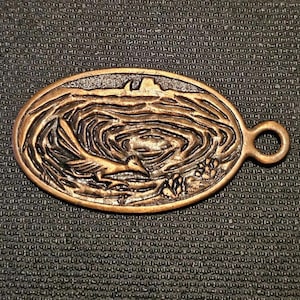 Könnte beinhalten: Ovale Anhänger aus Bronze mit einem detaillierten Relief eines Roadrunners in einer Wüstenlandschaft. Der Anhänger hat eine Öse zur Befestigung und ist vor einem strukturierten Hintergrund platziert.