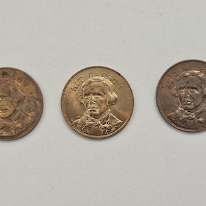 May include: Three bronze-colored commemorative coins. Each coin features a portrait of a man and the name "KIT CARSON" around the edge. The coins are arranged in a horizontal line against a white background.