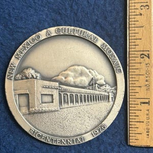 May include: A silver-colored commemorative coin with the text "NEW MEXICO A CULTURAL MOSAIC" and "BICENTENNIAL 1976". The coin features a building with a long colonnade, mountains, and a cloud. A wooden ruler is next to the coin.