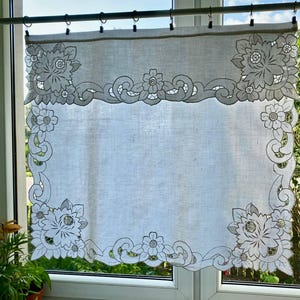 May include: A white window curtain featuring intricate cutwork embroidery with floral motifs along the borders and corners. The fabric is semi-sheer, and the curtain is suspended from a metal rod using small black clips attached to the top edge.