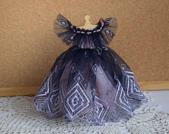 May include: A miniature doll dress with a layered design. The dress features a black tulle overlay with a geometric pattern over a pink underdress. The bodice has a decorative neckline and the dress is displayed on a wooden stand.