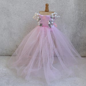 Peut inclure: Une robe miniature sur un mannequin en bois, dotée d'un corsage en satin rose clair avec des manches tombantes ornées de fausses perles blanches et de feuilles vertes, ainsi qu'une jupe volumineuse en tulle rose clair superposé.