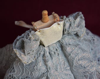 May include: A doll dress with a light blue lace overlay. The dress features a small, gold-coloured purse with beaded handles and a wooden peg doll head. The lace has a delicate floral pattern.