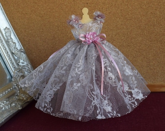 May include: A miniature dress made of delicate lace with a pink satin ribbon and a pink flower accent. The dress is displayed on a wooden mannequin.