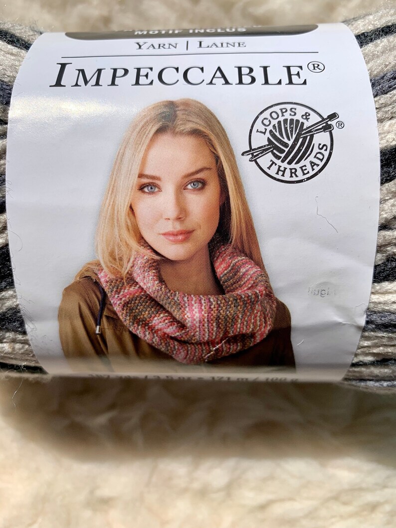 May include: A skein of Loops & Threads Impeccable yarn in a black, white, and beige colorway. The yarn is shown with a woman wearing a scarf made from the yarn.