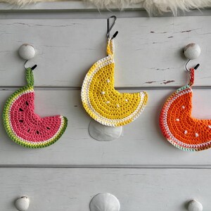 Puede incluir: Tres adornos de frutas de ganchillo colgados de ganchos. Los adornos tienen forma de sandía, limón y naranja. La sandía es rosa y verde, el limón es amarillo y blanco, y la naranja es naranja y blanca.
