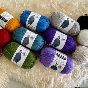 May include: A selection of multi-coloured yarn skeins, including shades of yellow, red, blue, green, purple, grey, black, and white. Each skein has a white label with text and an image. The yarn is arranged on a fluffy, cream-coloured surface.