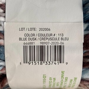 K&ouml;nnte beinhalten: Eine Garnrolle in Blau, Braun und Wei&szlig; mit dem Etikett "BLUE DUSK / CR&Eacute;PUSCULE BLEU" und der Losnummer "202006". Das Etikett enth&auml;lt auch die Farbnummer "113" und den UPC-Barcode.
