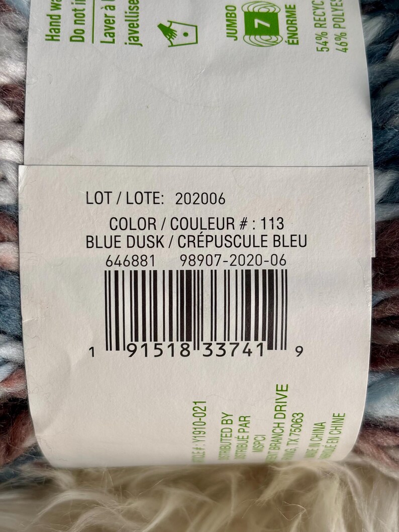 K&ouml;nnte beinhalten: Eine Garnrolle in Blau, Braun und Wei&szlig; mit dem Etikett "BLUE DUSK / CR&Eacute;PUSCULE BLEU" und der Losnummer "202006". Das Etikett enth&auml;lt auch die Farbnummer "113" und den UPC-Barcode.