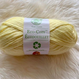 Peut inclure: Une pelote de laine jaune pâle, étiquetée "Eco-COZY Écoudouillet", avec un symbole de recyclage. La laine est fabriquée à partir de plastique recyclé et se trouve sur une surface blanche et moelleuse. L'étiquette indique également "Yarn | Laine".