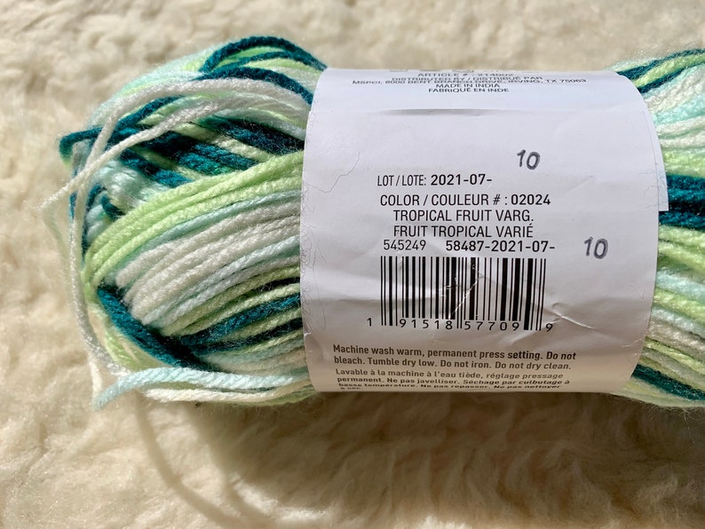 May include: A skein of yarn in a tropical fruit colorway. The yarn is white, green, and teal. The label reads "Tropical Fruit Varg. Fruit Tropical Vari&eacute;" and includes the lot number 2021-07- and color number 02024.