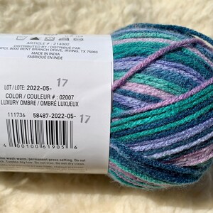 May include: A ball of yarn with a label that reads "LOT/LOTE: 2022-05-17 COLOR/COULEUR #: 02007 LUXURY OMBRE/OMBR&Eacute; LUXUEUX 111736 58487-2022-05-17" The yarn is a mix of blue, green, and purple shades.