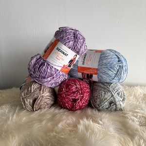 May include: Five balls of yarn in various colors, including purple, blue, pink, beige, and green. The yarn is labeled "Bernat Softee Chunky Twist".