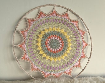 Puede incluir: Un gran tapiz de pared circular de ganchillo con un diseño de mandala. El mandala presenta una variedad de colores, incluyendo rosa, amarillo, verde y blanco. La pieza de ganchillo está unida a un aro de madera.