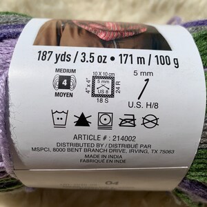 May include: A skein of yarn in shades of purple, green, and gray. The label reads "187 yds / 3.5 oz &bull; 171 m / 100 g" and includes care instructions and the article number "214002".