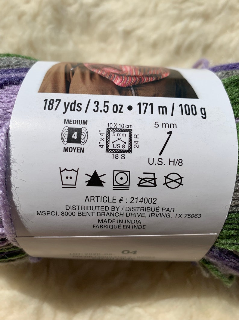 May include: A skein of yarn in shades of purple, green, and gray. The label reads "187 yds / 3.5 oz &bull; 171 m / 100 g" and includes care instructions and the article number "214002".