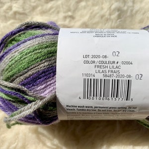 May include: A skein of yarn in shades of purple, green, and gray. The label reads "LOT: 2020-08-02", "COLOR/COULEUR #: 02004", "FRESH LILAC", "LILAS FRAIS", "110314", "58487-2020-08-02". Machine wash warm, permanent press setting. Do not bleach. Tumble dry low. Do not iron. Do not dry clean.