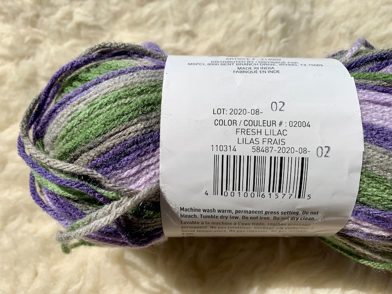 May include: A skein of yarn in shades of purple, green, and gray. The label reads "LOT: 2020-08-02", "COLOR/COULEUR #: 02004", "FRESH LILAC", "LILAS FRAIS", "110314", "58487-2020-08-02". Machine wash warm, permanent press setting. Do not bleach. Tumble dry low. Do not iron. Do not dry clean.