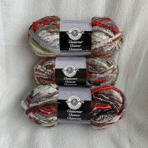 May include: Three skeins of Loops & Threads Glamorous Glamour yarn in a multi-coloured pattern with red, white, green, and brown shades. Each skein is 53 yards/49 metres.