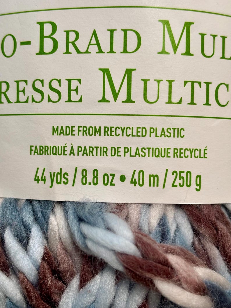 K&ouml;nnte beinhalten: Eine Kn&auml;uel dickes, mehrfarbiges Garn in Blaut&ouml;nen, Wei&szlig; und Braun. Das Garn besteht aus recyceltem Kunststoff und ist mit "O-BRAID MULTRESSE MULTIC" und dem Text "MADE FROM RECYCLED PLASTIC FABRIQU&Eacute; &Agrave; PARTIR DE PLASTIQUE RECYCL&Eacute; 44 yds / 8.8 oz &bull; 40 m / 250 g" beschriftet.