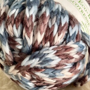 K&ouml;nnte beinhalten: Ein Kn&auml;uel Garn in Blaut&ouml;nen, Braun, Wei&szlig; und Rosa. Das Garn ist weich und dick und perfekt zum Stricken oder H&auml;keln.