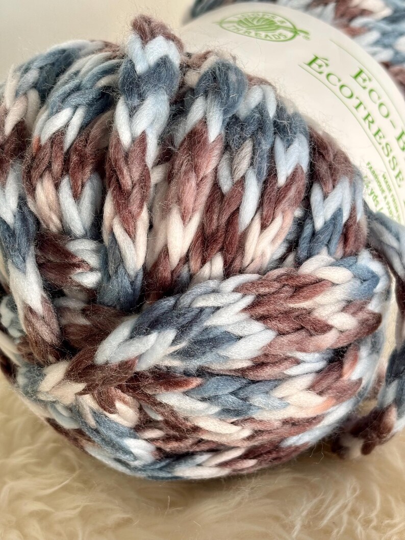 K&ouml;nnte beinhalten: Ein Kn&auml;uel Garn in Blaut&ouml;nen, Braun, Wei&szlig; und Rosa. Das Garn ist weich und dick und perfekt zum Stricken oder H&auml;keln.
