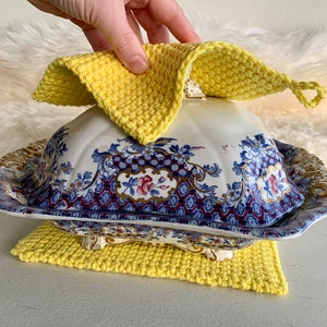 May include: A yellow crocheted dishcloth is placed on top of a vintage porcelain serving dish with a blue and gold floral pattern. The dish is sitting on a yellow crocheted coaster.