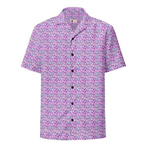 Puede incluir: Una camisa de manga corta con botones y un patrón repetitivo de formas irregulares y redondeadas en tonos morado, lavanda y azul claro. La camisa tiene un cuello clásico, una tapeta frontal con botones negros y un dobladillo recto.