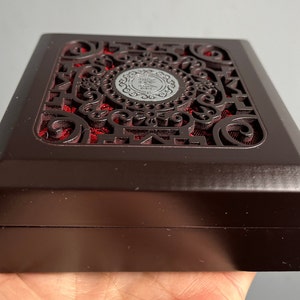 Könnte beinhalten: Eine dunkelbraune Holzkiste mit filigranen Ausschnitten und einem roten Samt-Innenraum. Die Kiste hat eine silberne Metallplakette mit arabischer Schrift.