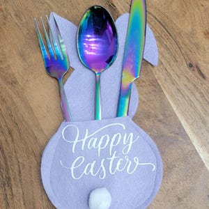 May include: A purple felt bunny-shaped utensil holder with the text "Happy Easter" in white. The holder has a white pom-pom for a tail and holds a rainbow-colored fork, spoon, and knife.