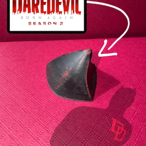 Puede incluir: Un accesorio negro y rojo de la serie Marvel Daredevil. El accesorio tiene una forma curva con una superficie texturizada. La imagen también incluye el logotipo de Daredevil y el texto "Born Again Temporada 2".