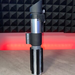 May include: A silver and black lightsaber prop with a rectangular viewing scope. The hilt has a ribbed design with black accents. The prop is made of plastic and metal and sits on a gray surface.